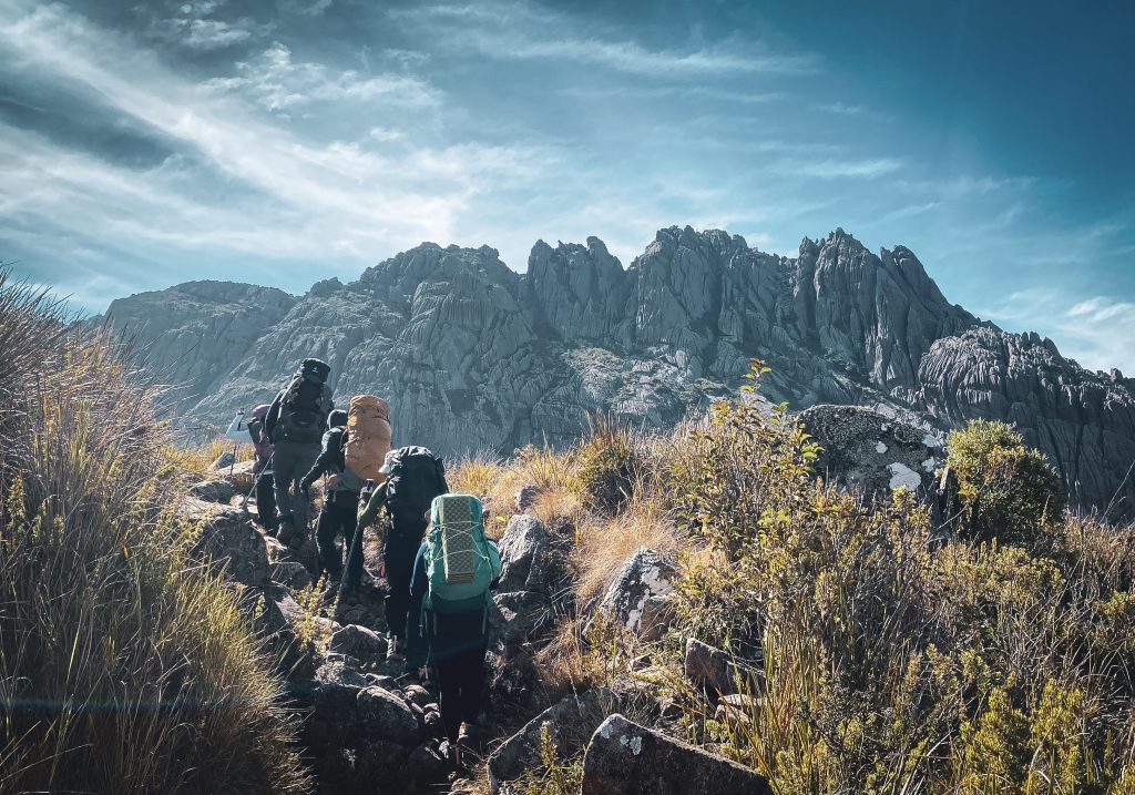 Grupo em trilha de terreno rochoso e inclinado com mochilas e bastões de caminhada.