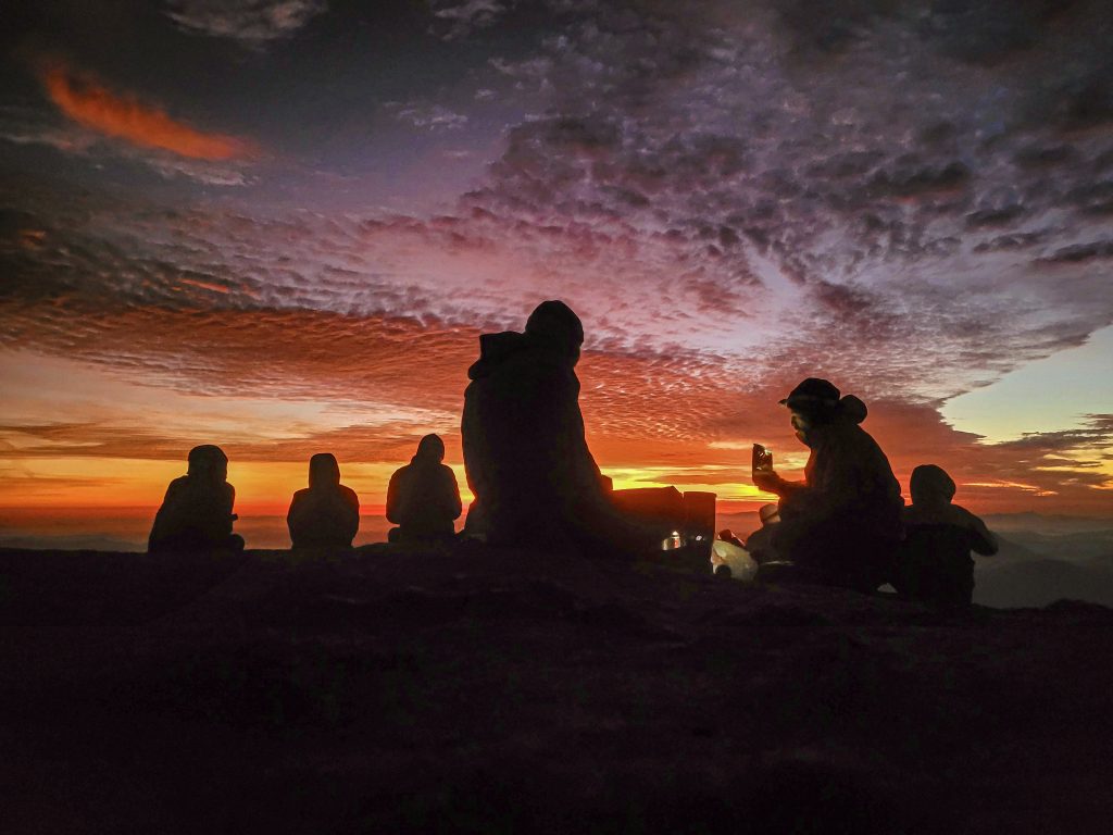Trekker chegando ao destino no fim do dia com mochila e bastões ao pôr do sol.