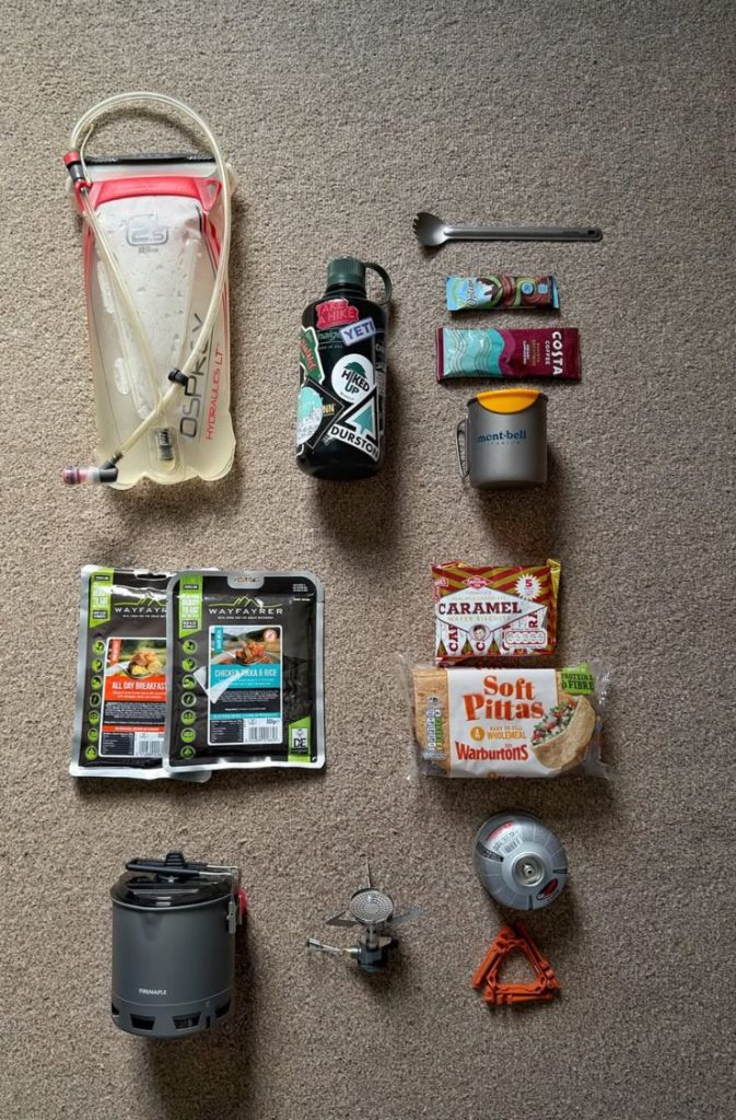Equipamentos essenciais organizados para trilha: mochila, lanterna, água, corta-vento, primeiros socorros e mapa.